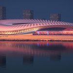 Photorealistic render of a stadium design with a large expanse of water in the foreground. The stadium is white with a large curved roof.