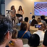 Two women standing in front of a lectern with a large display screen behind them, they are giving a presentation to a seated audience.