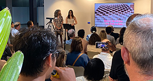 Two women standing in front of a lectern with a large display screen behind them, they are giving a presentation to a seated audience.