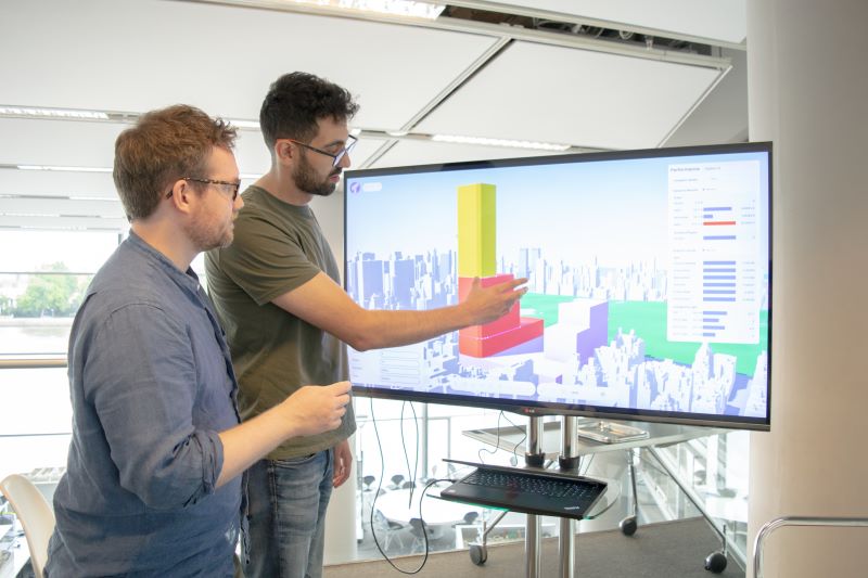 Image shows two people standing and looking to a large screen which shows a computer generated outline of buildings and a skyline.